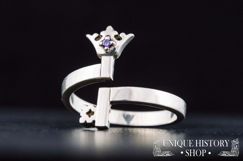 May include: A silver ring shaped like a key with a small purple gemstone on the crown. The ring is open and adjustable.