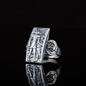 May include: A silver ring with a rectangular design featuring an engraved figure and a small red gemstone. The ring is set on a black background.