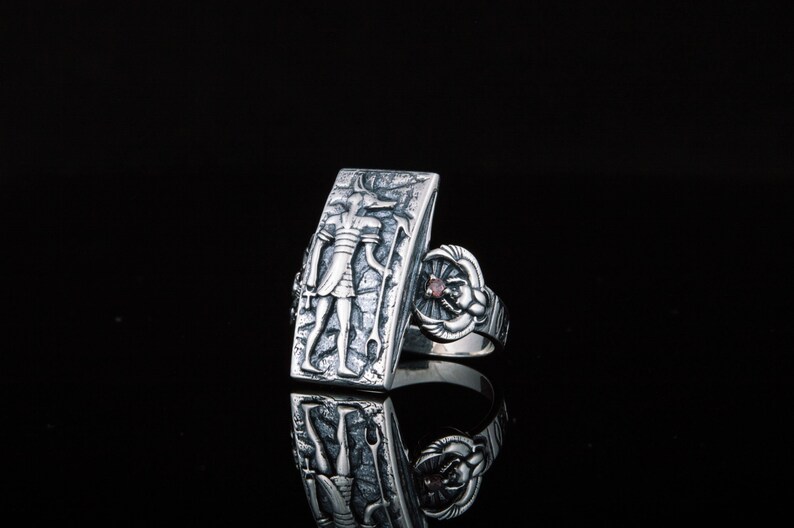 May include: A silver ring with a rectangular design featuring an engraved figure and a small red gemstone. The ring is set on a black background.