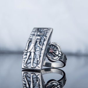 May include: A silver ring with a rectangular design featuring an engraved image of an Egyptian god. The ring has a small red gemstone on the side.