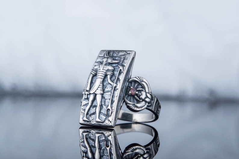 May include: A silver ring with a rectangular design featuring an engraved image of an Egyptian god. The ring has a small red gemstone on the side.