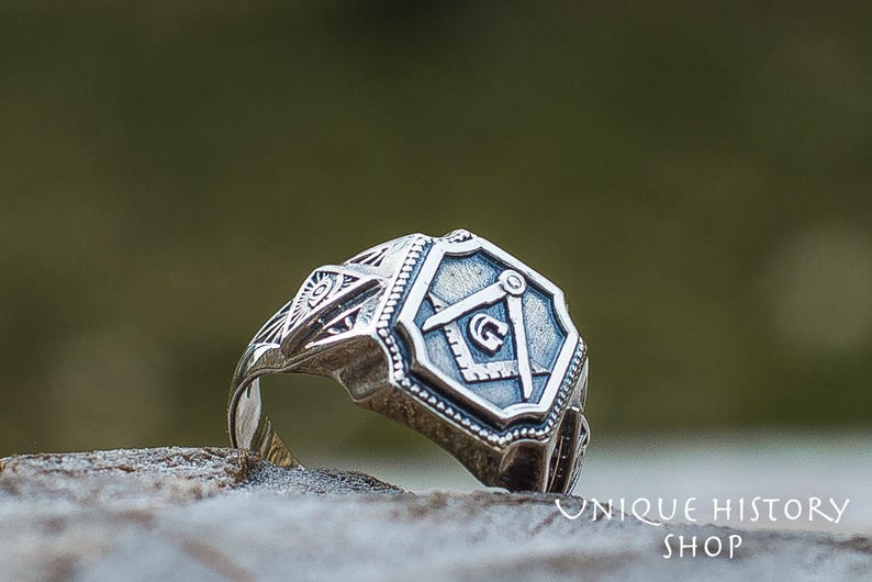 K&ouml;nnte beinhalten: Ein silberner Ring mit einem freimaurerischen Symbol, einem Zirkel und einem Winkelmesser, auf der Vorderseite. Der Ring ist mit komplizierten Designs verziert und liegt auf einem grauen Stein.