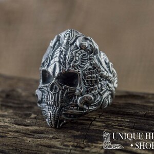 Peut inclure: Une bague en argent d&eacute;taill&eacute;e repr&eacute;sentant un cr&acirc;ne avec des gravures complexes. La bague pr&eacute;sente un design de cr&acirc;ne avec des traits faciaux d&eacute;taill&eacute;s et des ornements. La bague est pr&eacute;sent&eacute;e sur une surface en bois, avec le texte "UNIQUE HISTORY SHOP".