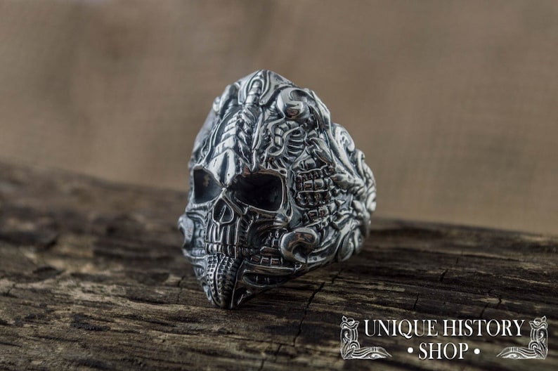 Peut inclure: Une bague en argent d&eacute;taill&eacute;e repr&eacute;sentant un cr&acirc;ne avec des gravures complexes. La bague pr&eacute;sente un design de cr&acirc;ne avec des traits faciaux d&eacute;taill&eacute;s et des ornements. La bague est pr&eacute;sent&eacute;e sur une surface en bois, avec le texte "UNIQUE HISTORY SHOP".