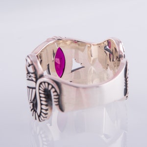 May include: Silver ring with an ornate design and a large, oval-shaped, magenta gemstone. The ring has detailed carvings and a polished finish. The ring is set against a white background.