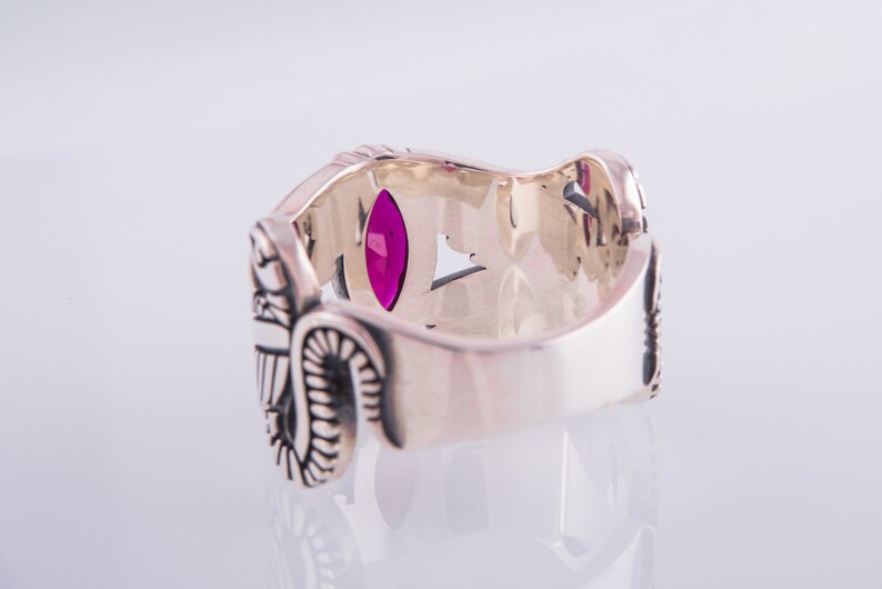 May include: Silver ring with an ornate design and a large, oval-shaped, magenta gemstone. The ring has detailed carvings and a polished finish. The ring is set against a white background.