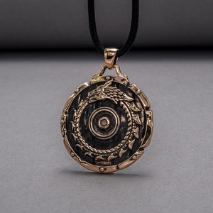 Pode incluir: Um pingente redondo de bronze com um design detalhado de um drag&atilde;o enrolado em torno de um c&iacute;rculo. O pingente tem uma pequena gema rosa no centro.