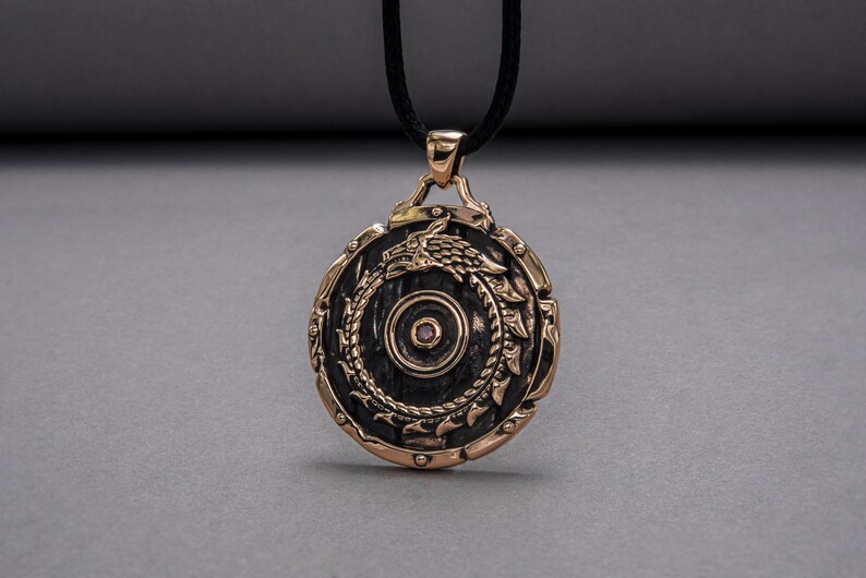 Pode incluir: Um pingente redondo de bronze com um design detalhado de um drag&atilde;o enrolado em torno de um c&iacute;rculo. O pingente tem uma pequena gema rosa no centro.