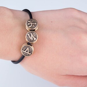 May include: A black elastic bracelet with three gold-colored beads. Each bead features a different rune symbol.