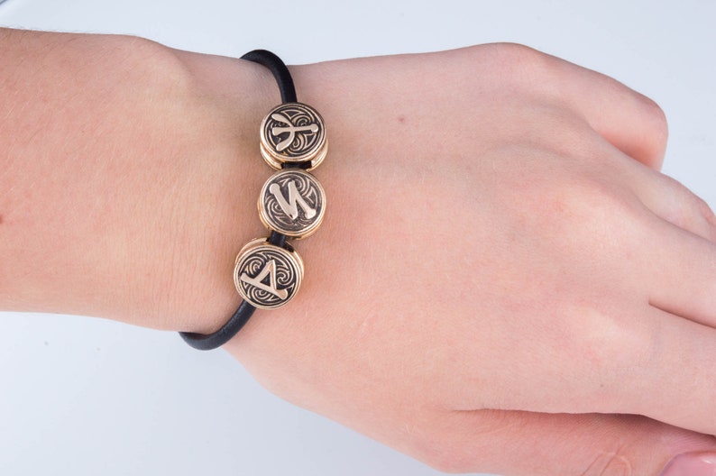 May include: A black elastic bracelet with three gold-colored beads. Each bead features a different rune symbol.