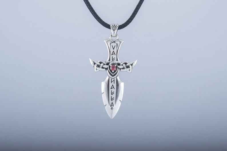 May include: Silver pendant necklace featuring a sword design with a red gemstone. The sword has the word "VALHALLA" inscribed on it.