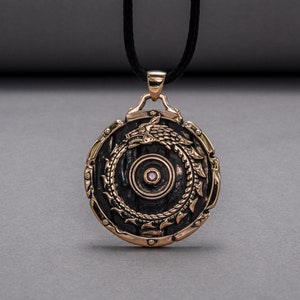 Pode incluir: Um pingente redondo de bronze com um centro preto e um design detalhado de um drag&atilde;o envolto em torno do centro. O pingente tem uma pequena gema rosa no centro.