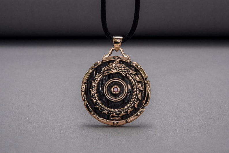 Pode incluir: Um pingente redondo de bronze com um centro preto e um design detalhado de um drag&atilde;o envolto em torno do centro. O pingente tem uma pequena gema rosa no centro.