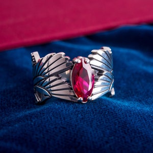 May include: A silver ring with a marquise-cut, ruby-red gemstone. The ring has a detailed, openwork design with stylized feather or leaf motifs. The ring is set against a deep blue background.