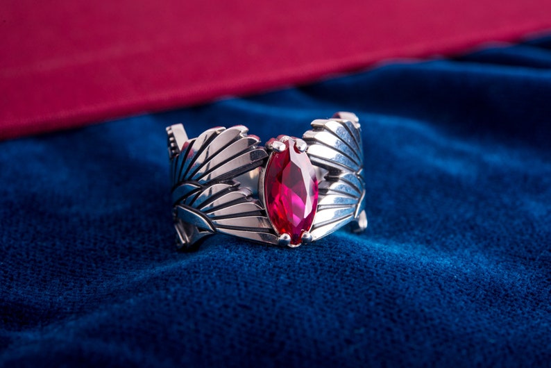 May include: A silver ring with a marquise-cut, ruby-red gemstone. The ring has a detailed, openwork design with stylized feather or leaf motifs. The ring is set against a deep blue background.