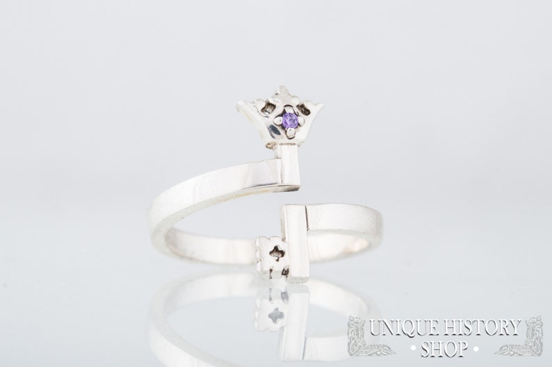 May include: A silver ring with a key design. The key has a small purple gemstone at the top, which is shaped like a crown.