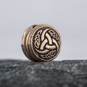 May include: A round, bronze-colored pendant with a Celtic knot design featuring a triquetra symbol. The pendant is on a gray rock.