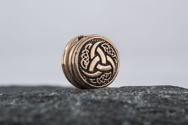 May include: A round, bronze-colored pendant with a Celtic knot design featuring a triquetra symbol. The pendant is on a gray rock.