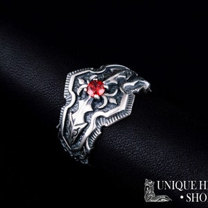 May include: A silver ring with an ornate shield design and a central red gemstone. The ring features intricate detailing and a dark background, highlighting the jewelry's craftsmanship. The text "UNIQUE HISTORY SHOP" is visible.