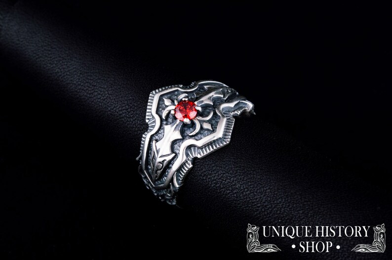 May include: A silver ring with an ornate shield design and a central red gemstone. The ring features intricate detailing and a dark background, highlighting the jewelry's craftsmanship. The text "UNIQUE HISTORY SHOP" is visible.
