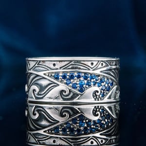 Può includere: Un anello d'argento con un design a onde e pietre preziose blu. L'anello è posizionato su uno sfondo blu scuro.