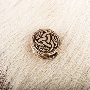 May include: A round, gold-colored metal pendant with a Celtic knot design featuring a triskelion symbol. The pendant is on a white furry surface.