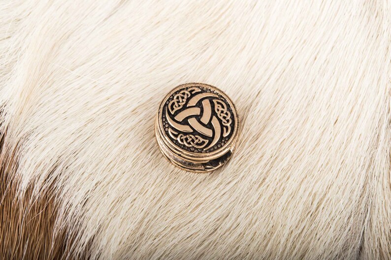 May include: A round, gold-colored metal pendant with a Celtic knot design featuring a triskelion symbol. The pendant is on a white furry surface.