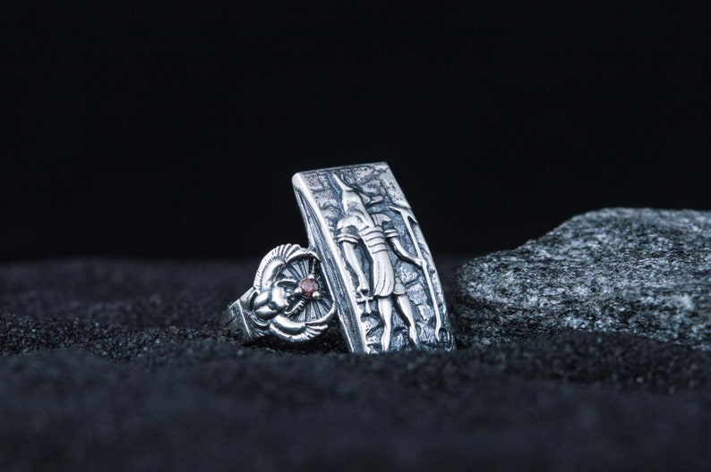 May include: Two silver rings with intricate designs. One ring features a detailed depiction of an Egyptian god, while the other ring has a red gemstone.