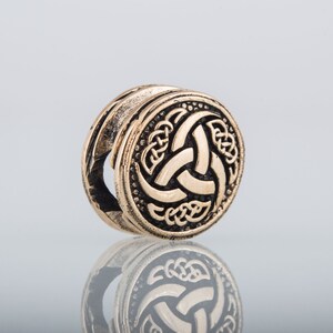 May include: A gold-colored metal bead with a black and gold design of a triskelion, a three-legged spiral, surrounded by a Celtic knot pattern.