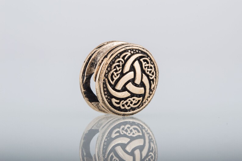 May include: A gold-colored metal bead with a black and gold design of a triskelion, a three-legged spiral, surrounded by a Celtic knot pattern.