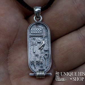 May include: Silver pendant featuring an Egyptian cartouche with hieroglyphs, including an ankh, a bird, and a staff. The pendant is on a black cord.