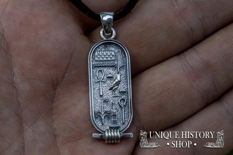 May include: Silver pendant featuring an Egyptian cartouche with hieroglyphs, including an ankh, a bird, and a staff. The pendant is on a black cord.