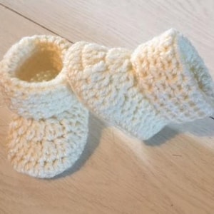 Puede incluir: Un par de botitas de bebé de ganchillo en color crema. Las botitas están hechas con un patrón de punto sencillo y tienen un aspecto suave y texturizado.