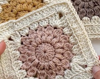 May include: Close-up of three crocheted granny squares in cream, tan, and mauve. The squares feature a floral center design and are held by a person's hands. The crochet work is detailed and textured, showcasing the craft.