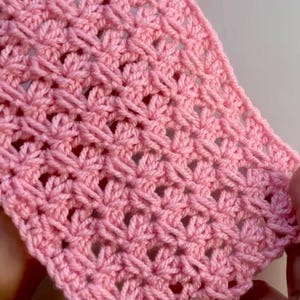 Könnte beinhalten: Nahaufnahme eines Abschnitts aus rosa Häkelstoff. Das Stichmuster erzeugt ein strukturiertes, offenes Design mit blütenartigem Aussehen. Das weiche rosa Garn ist eng gewebt und zeigt die detaillierte Handwerkskunst des handgefertigten Artikels.