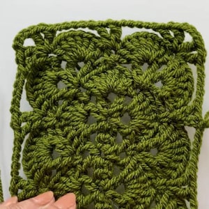 May include: Close-up of a square, olive green crochet piece. The handmade item features a detailed, openwork design with a scalloped edge. The yarn is a solid, dark green color, and the texture is clearly visible.