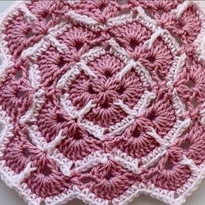 May include: A handmade, square crochet doily in shades of pink and white. The design features a repeating pattern of fan-shaped motifs and a scalloped edge. The doily is approximately 20 cm wide.