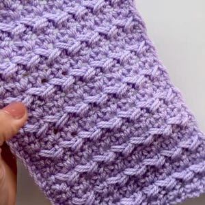 Pode incluir: Close-up de uma seção de tecido de crochê em uma cor lavanda suave. O padrão de ponto cria uma aparência texturizada e tecida. O fio é bem tecido, mostrando o artesanato detalhado do crochê. O tecido é segurado por uma mão.