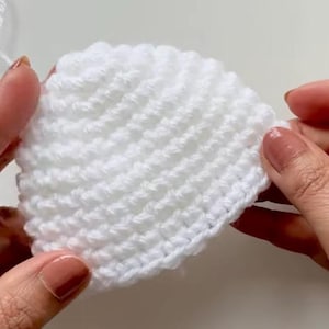 May include: A white, crocheted, semi-circular piece of fabric. The close-up shot shows the texture of the yarn and the stitches. The piece is held between two hands, with the fingers and nails visible.