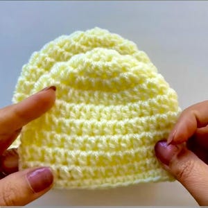 May include: A handmade, pale yellow crochet hat. The hat is held by two hands, showcasing the textured stitch pattern. The hat's shape is rounded, with a slightly scalloped edge. The yarn appears soft and the color is a light, pastel yellow.