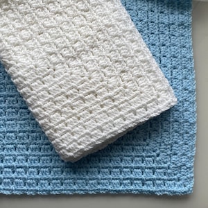 May include: A white crocheted blanket folded over a light blue crocheted blanket. The blankets have a textured, woven appearance. The white blanket is folded at an angle, partially covering the blue blanket. The image is well-lit, highlighting the texture and color of the blankets.