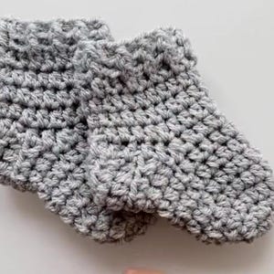 May include: A pair of handmade, grey crocheted baby booties. The booties are made with a thick yarn, and have a textured appearance. The cuffs are folded over. The booties are designed to keep a baby's feet warm.