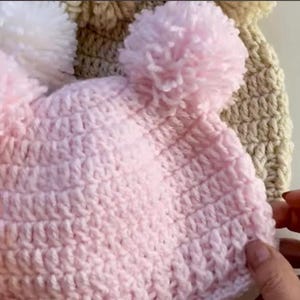 May include: Three crocheted hats with pom-poms. One hat is pink with two white pom-poms, one is beige with a white pom-pom, and one is white with a white pom-pom.