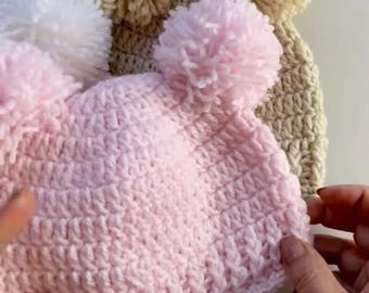 Puede incluir: Tres gorros de punto con pompones. Un gorro es rosa con dos pompones blancos, uno es beige con un pompón blanco y uno es blanco con un pompón blanco.