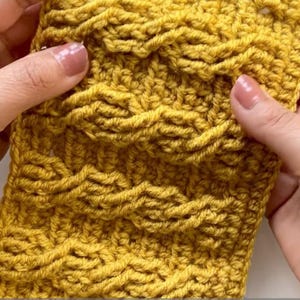 May include: Close-up of a mustard yellow crochet swatch with a cable stitch pattern. The textured fabric is held between two hands, showcasing the detailed craftsmanship. The yarn appears soft and the stitches are well-defined.