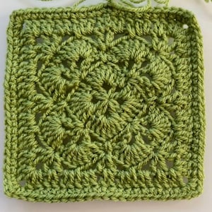 May include: A square, handmade crochet piece in shades of green. The intricate design features a central floral motif surrounded by detailed patterns. The edges are neatly finished, and loose yarn strands are visible.