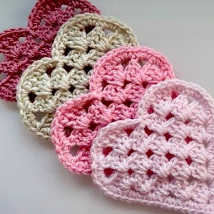 Op de afbeelding: Een verzameling van vijf gehaakte hartvormige items in verschillende tinten roze en crème. De handgemaakte harten zijn diagonaal gerangschikt en tonen het open, getextureerde steekpatroon. Het kleurenpalet omvat licht- en donkerroze en een crèmekleur.