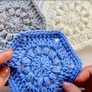 May include: Close-up of three crocheted squares in shades of blue, grey, and white. The blue square is in focus, held by hands, showcasing a detailed floral pattern. The other squares are in the background, with a similar design.