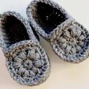 May include: A pair of grey crocheted slippers with a flower design. The slippers are made with a soft yarn and have a comfortable fit.