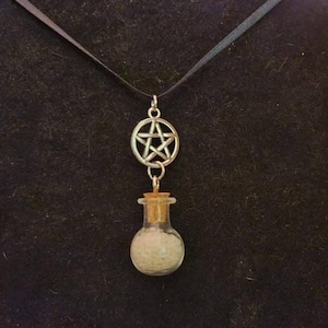 Peut inclure: Un collier à ruban noir avec un pendentif pentagramme argenté et un petit flacon en verre rempli de granules blancs. Le flacon est bouché avec un bouchon en liège et est suspendu sous le pentagramme.